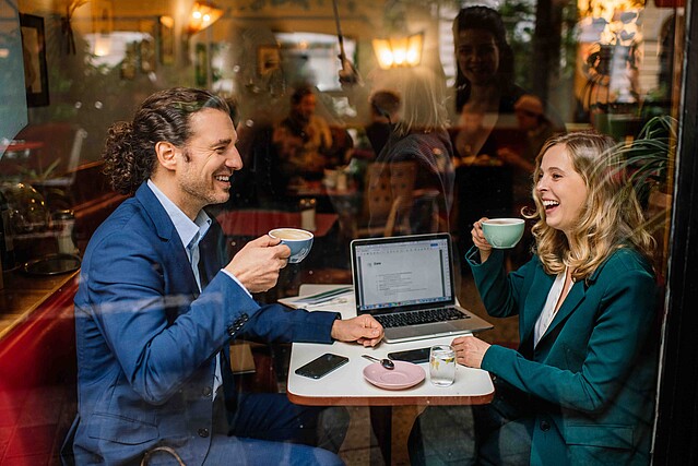 A man and a woman in business attire are seen through a coffee shop window sipping drinks and laughing with a laptop behind them.