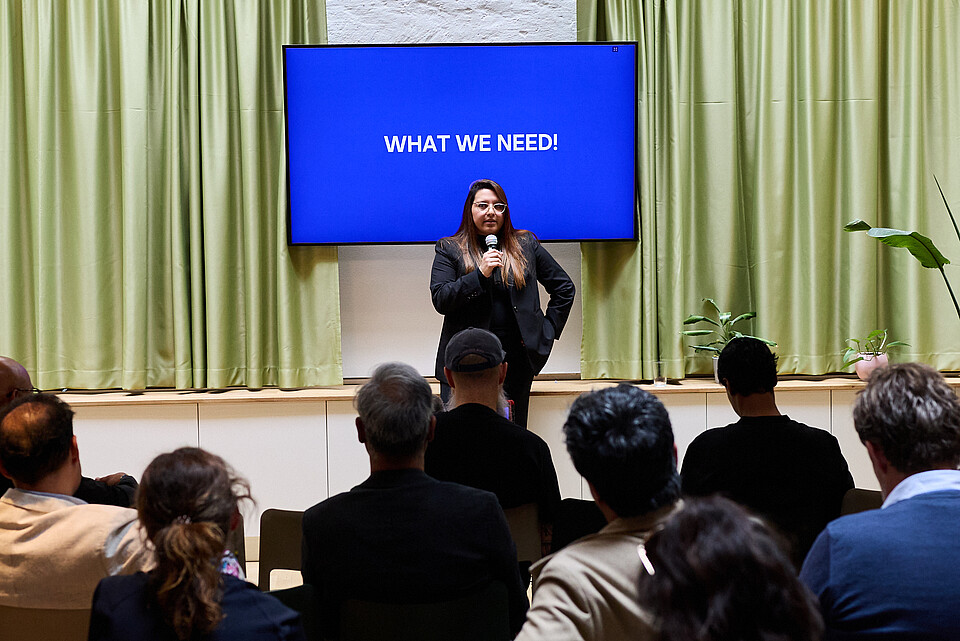 Woman with a microphone standing in front of a blue screen which reads 'What we need!'