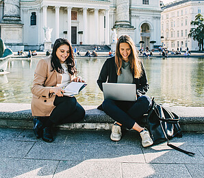 Two women working on the laptop on the edge of a fountain in front of Vienna's Karlskirche.