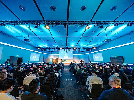 A view from the back of a conference room filled with people, and people pitching on stage.