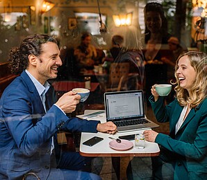 A man and a woman in business attire are seen through a coffee shop window sipping drinks and laughing with a laptop behind them.