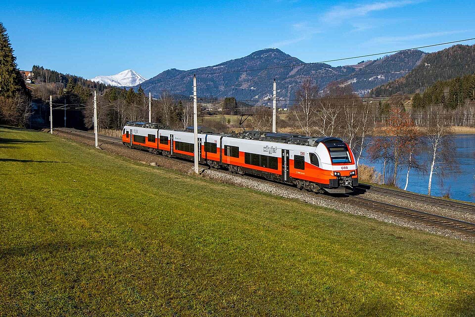 A red and white OBB train driving along a lake in the mountains