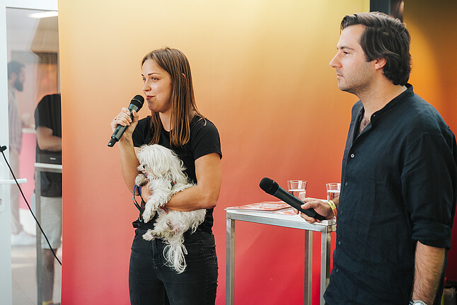 A woman, a man, and a dog, on stage at an event, speaking into the microphone.