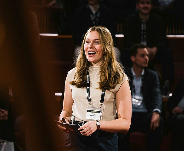 A woman with a lanyard and prompt cards speaking in front of a crowd